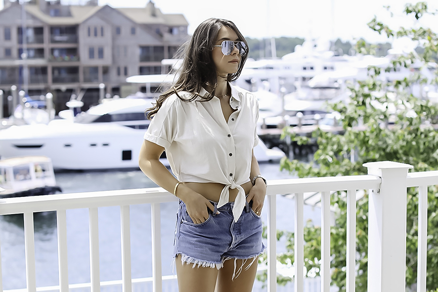 Liv on a deck overlooking the wharf in Newport Rhode Island wearing Urban Outfitter shirt, vintage Levi jeans by Re Done, and Quay Key Sunglasses